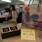 辦公室桌上擺放著來自日本的伴手禮，兩位女同事正在討論是否要將伴手禮分享給辦公室所有同仁。照片呈現文章中的真實情境，透過一場關於伴手禮的對話，引發對送禮文化、人際關係、心意、價值觀與每個人理解世界規則不同的深刻思考。